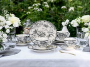 Schwarz-weißes Porzellan-Kaffeeservice mit floralen Motiven und Vogel-Illustration auf gedecktem Tisch im Garten, frontale Perspektive auf die Tassen und den Teller im Hintergrund