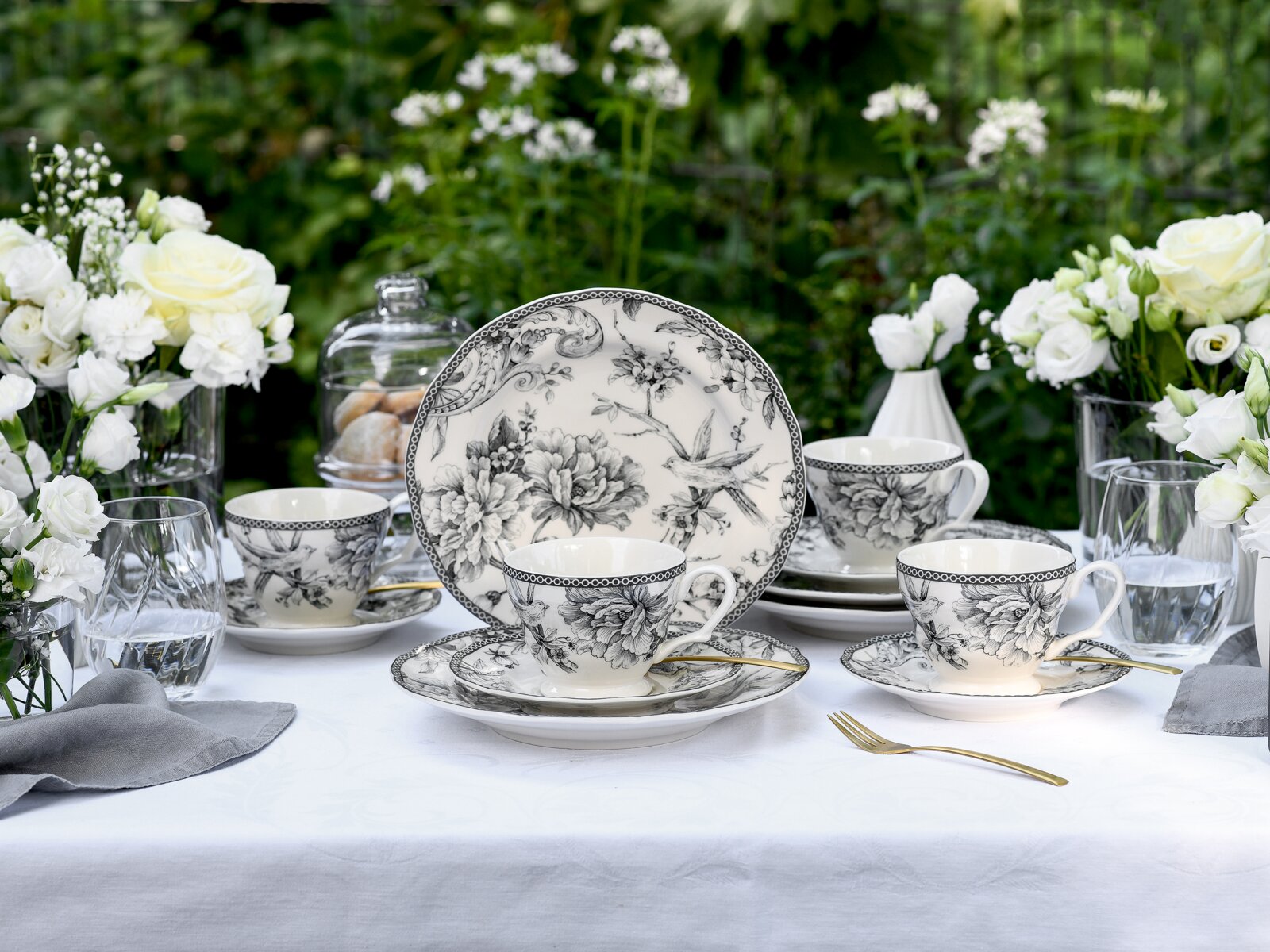 creaTable Kaffeeservice 12-tlg ADELAIDE Schwarz-weißes Porzellan-Kaffeeservice mit floralen Motiven und Vogel-Illustration auf gedecktem Tisch im Garten, frontale Perspektive auf die Tassen und den Teller im Hintergrund