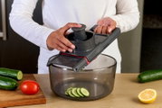 Hand hält grauen Gemüsehobel/Mandoline mit Fingerschutz über einer transparenten Schüssel, Gurkenscheiben fallen hinein; Perspektive: leicht von vorne/oben.