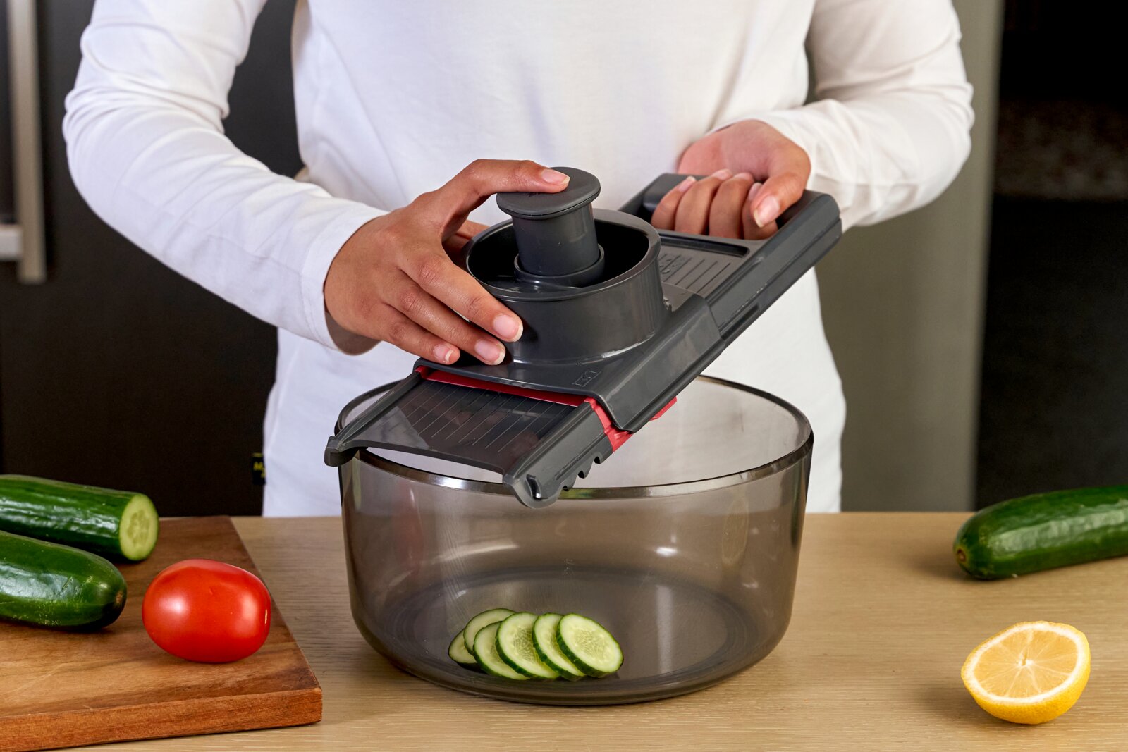 Hand hält grauen Gemüsehobel/Mandoline mit Fingerschutz über einer transparenten Schüssel, Gurkenscheiben fallen hinein; Perspektive: leicht von vorne/oben.