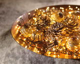 Nahaufnahme einer dekorativen LED-Clusterbeleuchtung auf einem goldenen Tablett, umgeben von Tannenzapfen und kleinen Glasornamenten.