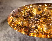 Nahaufnahme einer dekorativen LED-Clusterbeleuchtung auf einem goldenen Tablett, umgeben von Tannenzapfen und kleinen Glasornamenten.