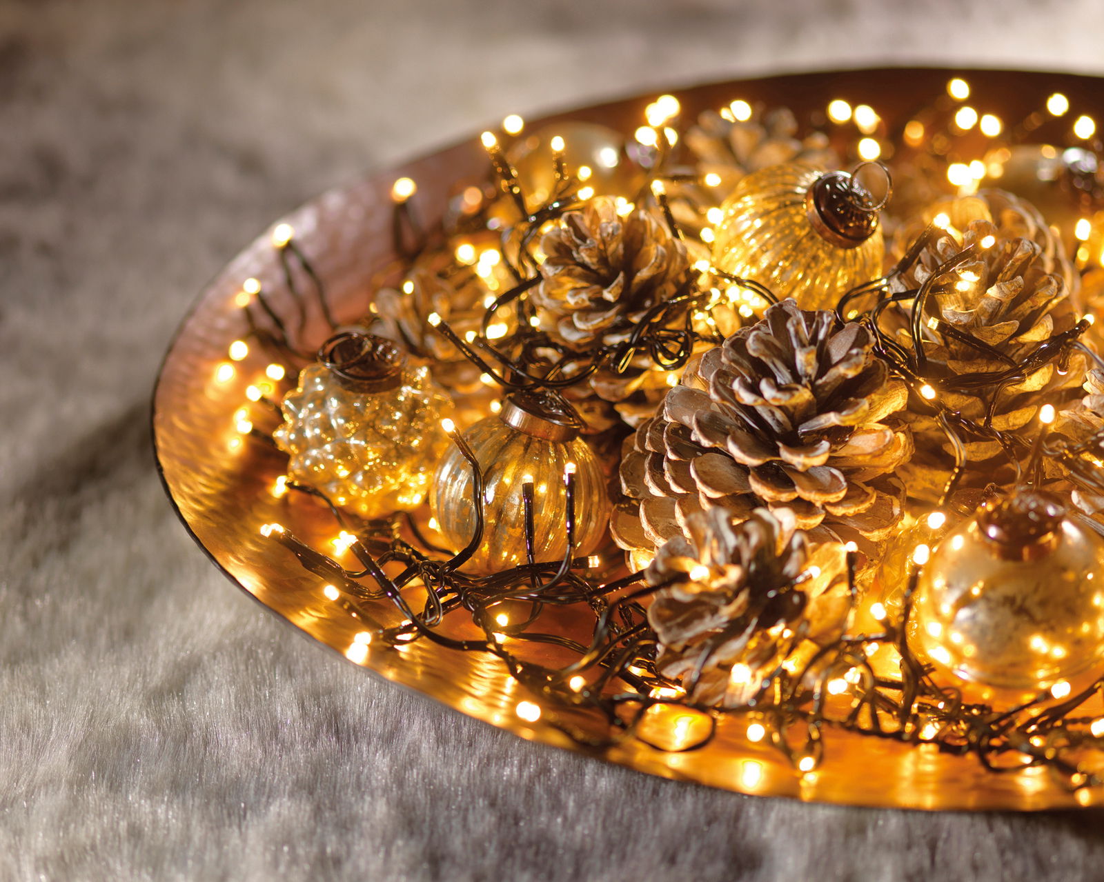 Nahaufnahme einer dekorativen LED-Clusterbeleuchtung auf einem goldenen Tablett, umgeben von Tannenzapfen und kleinen Glasornamenten.