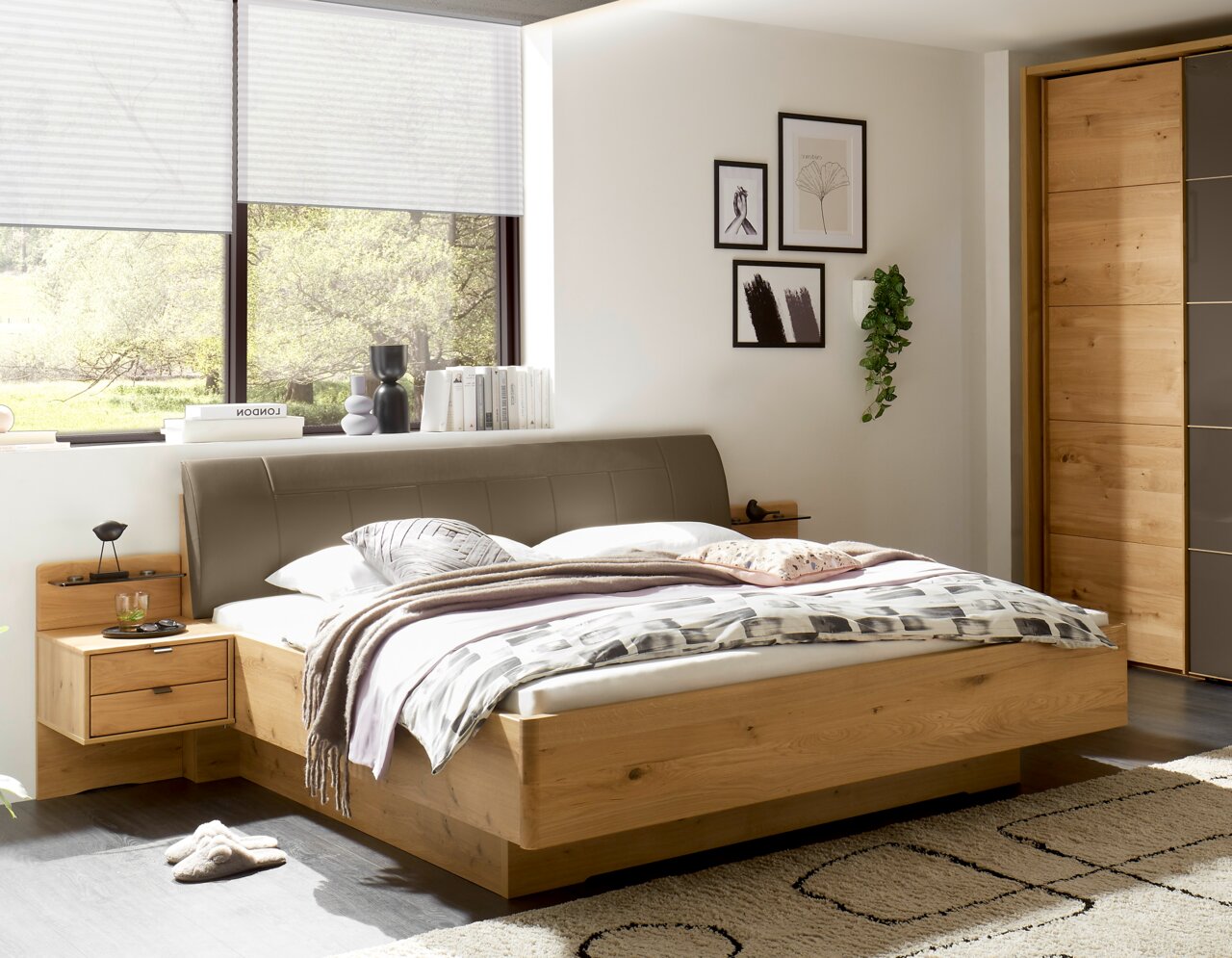 Modern houten bed met een grijs gestoffeerd hoofdeinde in een lichte slaapkamer, met nachtkastje en beddengoed; perspectief: vooraanzicht, licht van links.