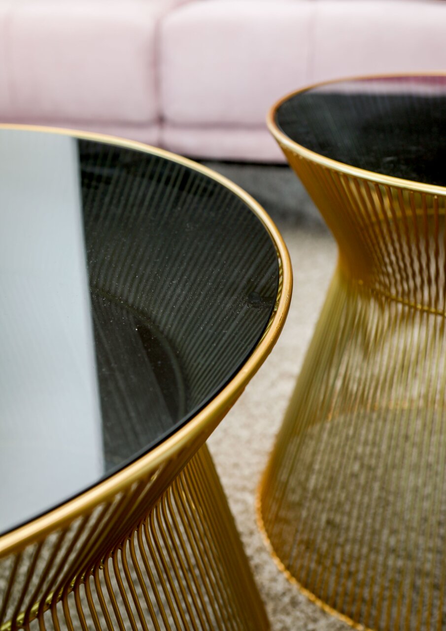 Close-up van twee elegante tafels met gouden metalen onderstellen en donkere glazen bladen, met op de achtergrond een roze bank.