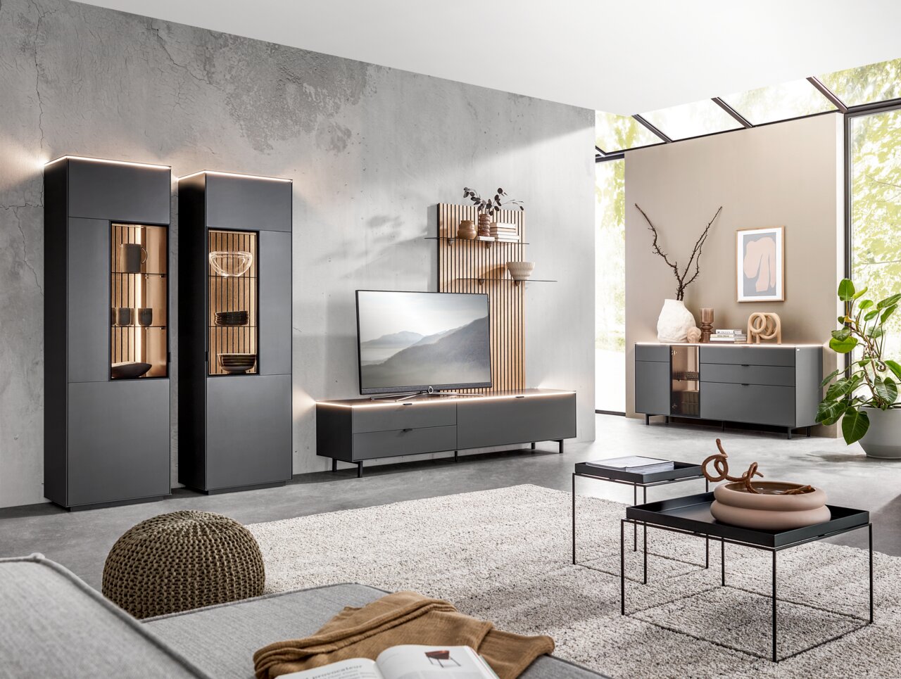 Moderne Wohnzimmermöbel in einem hellen Raum mit zwei hohen Vitrinen, einem TV-Lowboard und einem Sideboard, aus der Perspektive von vorne betrachtet.