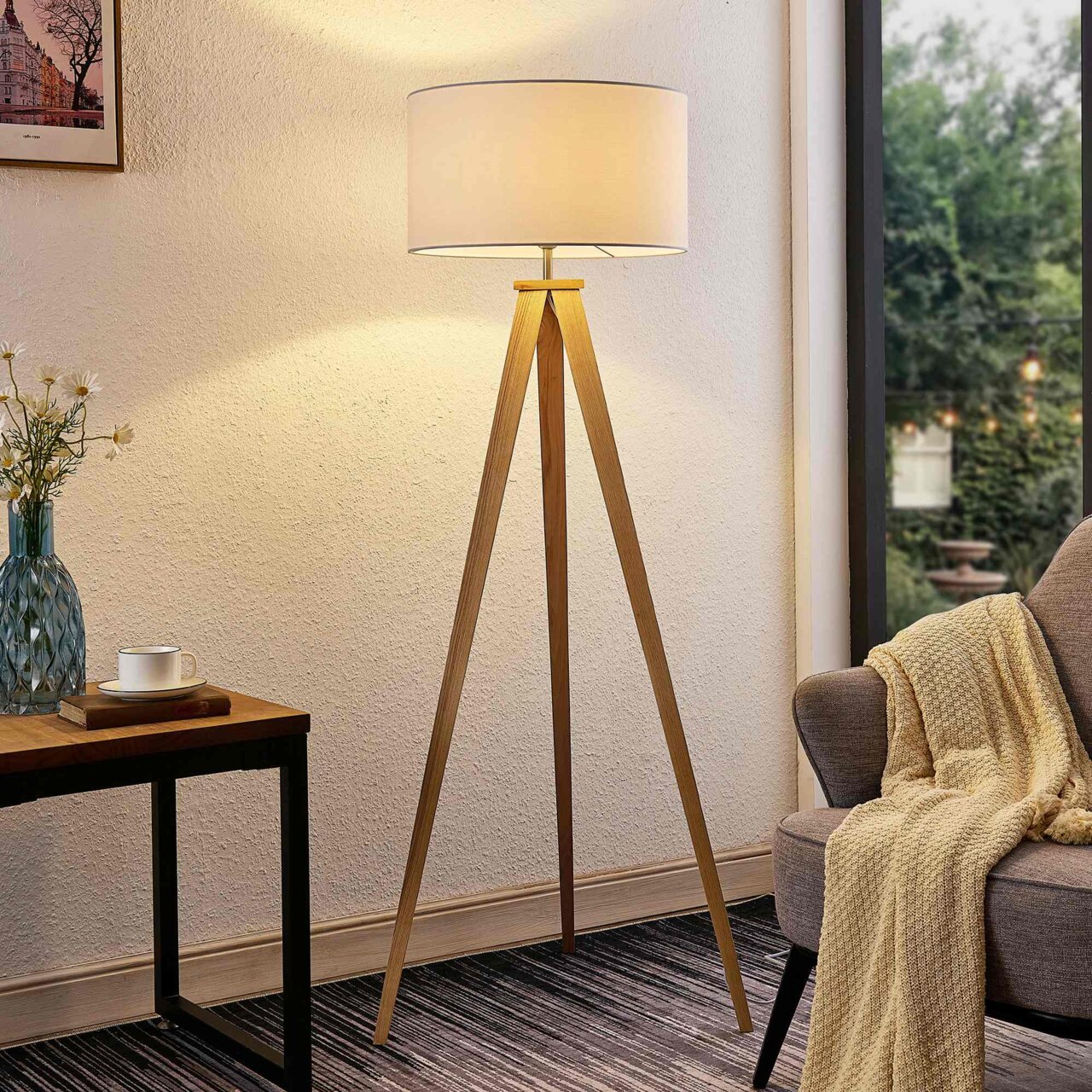 Moderne Stehleuchte mit dreibeinigem Holzgestell und rundem, weißem Lampenschirm, seitliche Perspektive im Wohnzimmer.