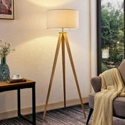 Moderne Stehleuchte mit dreibeinigem Holzgestell und rundem, weißem Lampenschirm, seitliche Perspektive im Wohnzimmer.