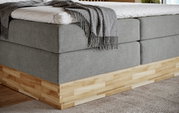 Eckansicht eines Boxspringbetts mit grauem Stoffbezug und Holzunterbau, darauf liegt eine graue Decke.