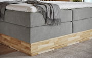 Eckansicht eines Boxspringbetts mit grauem Stoffbezug und Holzunterbau, darauf liegt eine graue Decke.
