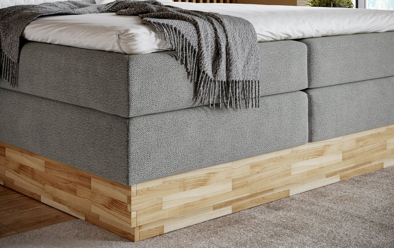 Zijaanzicht van een boxspringbed met een grijze stoffen bekleding en een houten onderstel, met daarop een grijze deken.