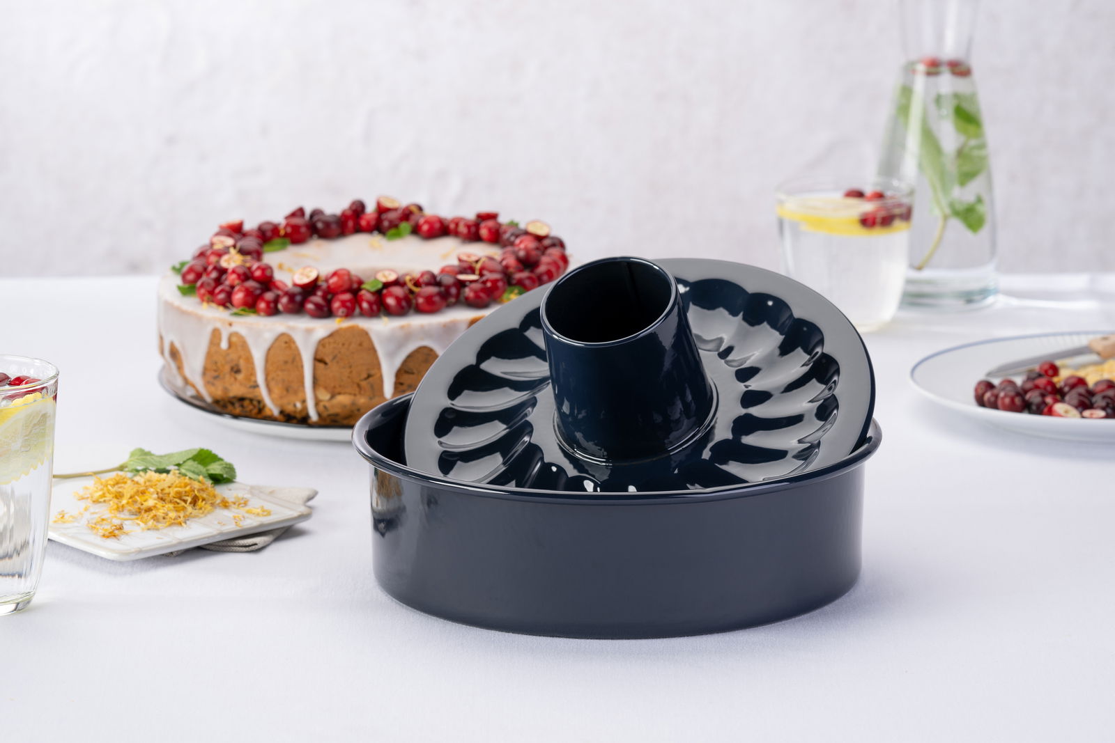 Schwarze Gugelhupfform aus Metall, seitliche Perspektive, auf einem Tisch mit einem dekorierten Kuchen im Hintergrund.