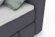 Close-up van een boxspringbed met grijze stoffen bekleding en een wit matras, gezien vanaf de zijkant