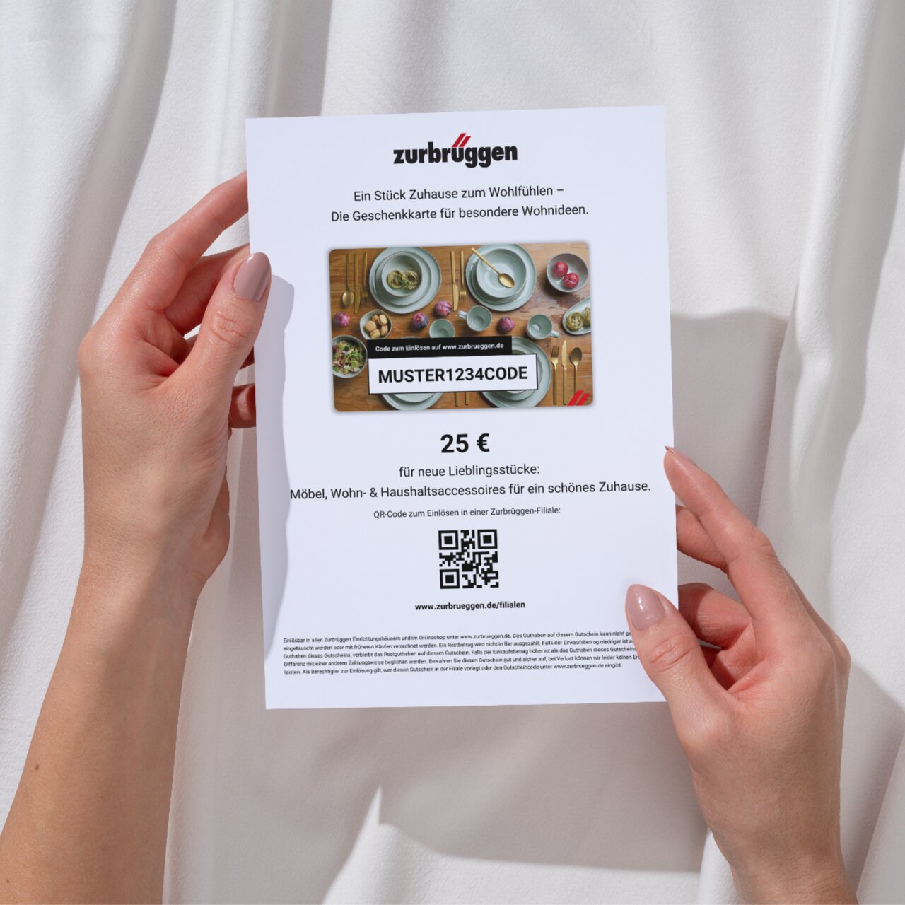 Zurbrüggen Digitaler Geschenkgutschein zum Ausdrucken Eine Person hält eine Zurbrüggen-Geschenkkarte im Wert von 25 Euro in den Händen, mit einem Bild von Geschirr und Besteck. Perspektive von oben auf die Karte.
