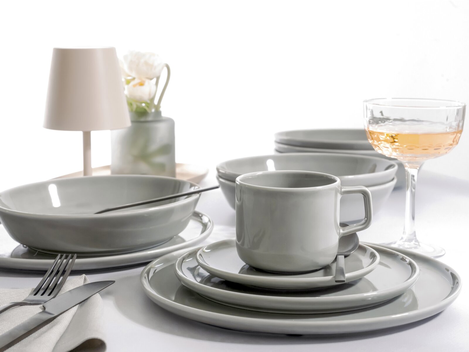 Graues Keramik-Geschirrset mit Tasse, Teller und Schalen auf gedecktem Tisch, seitliche Nahaufnahme in leicht erhöhter Perspektive; im Hintergrund Lampe, Vase und Glas mit Getränk