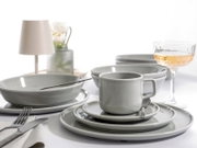 Graues Keramik-Geschirrset mit Tasse, Teller und Schalen auf gedecktem Tisch, seitliche Nahaufnahme in leicht erhöhter Perspektive; im Hintergrund Lampe, Vase und Glas mit Getränk