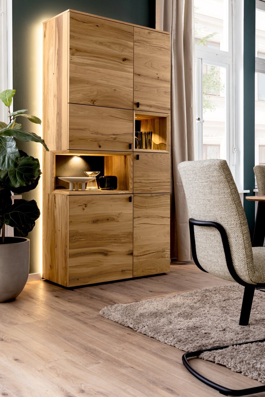 Venjakob Vitrine ANDIAMO 3.0 Hoher Wohnzimmerschrank aus hellem Massivholz mit mehreren Türen und offenem, beleuchtetem Regalfach, aufgenommen in schräger Frontansicht von links in einem modernen Wohnraum.