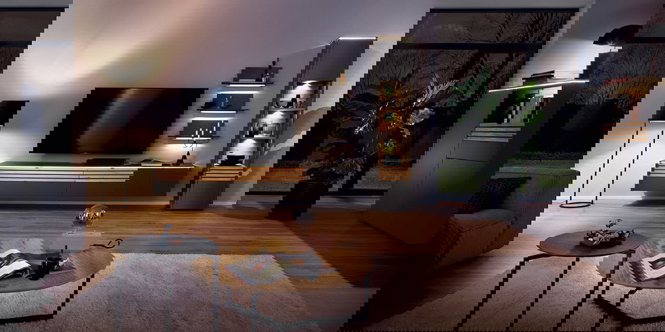 Ein modernes Wohnzimmer mit dunklen Möbeln, stimmungsvoller Beleuchtung, großem Fernseher und Pflanzen.