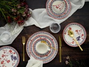 Draufsicht auf ein gedecktes Tischarrangement mit floral gemustertem Porzellan-Geschirr (Teller und Tassen) in Rot-Blau, goldfarbenem Besteck, Leinenserviette, Wassergläsern und Blumen auf dunklem Holztisch.