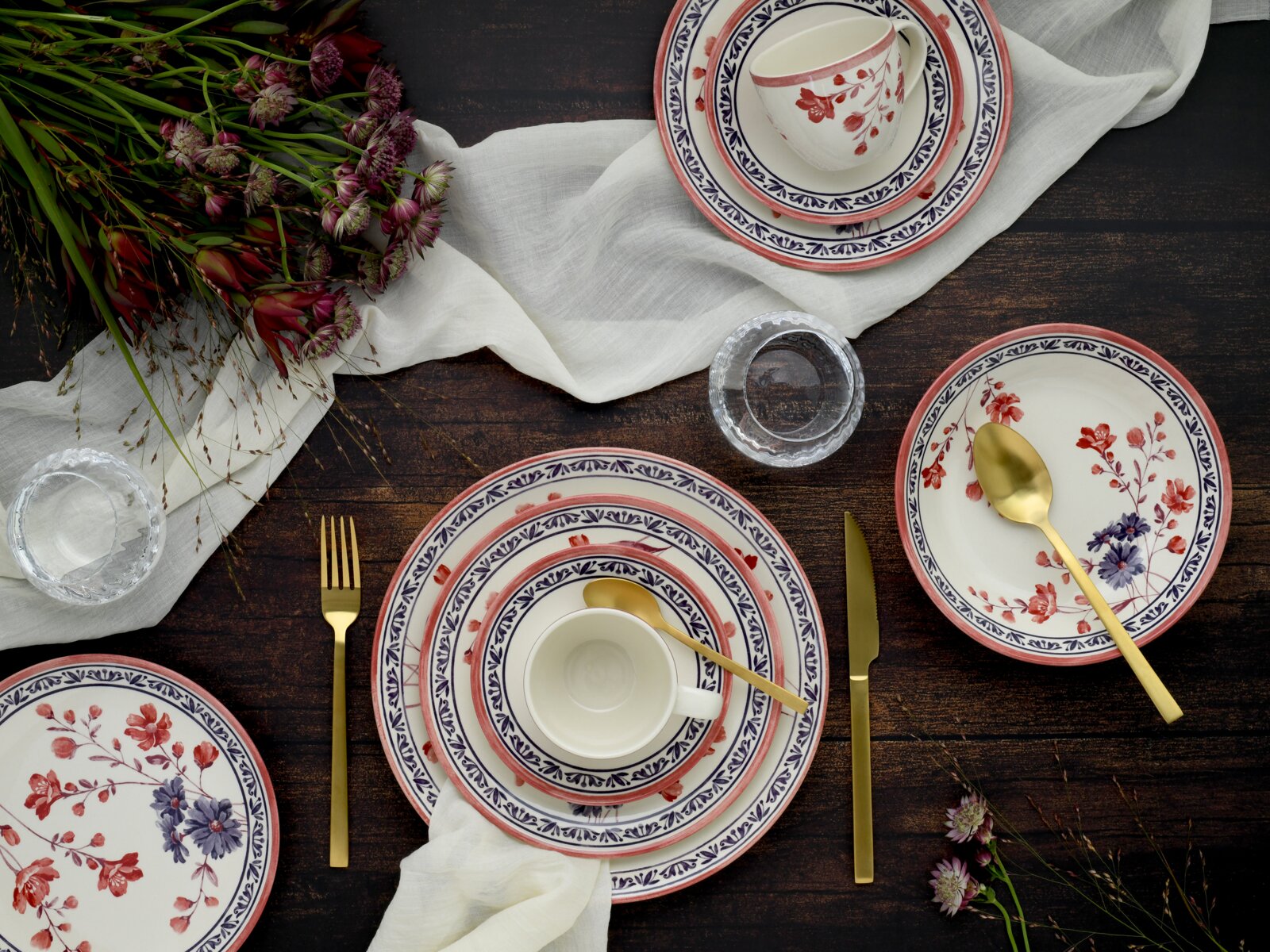 Draufsicht auf ein gedecktes Tischarrangement mit floral gemustertem Porzellan-Geschirr (Teller und Tassen) in Rot-Blau, goldfarbenem Besteck, Leinenserviette, Wassergläsern und Blumen auf dunklem Holztisch.
