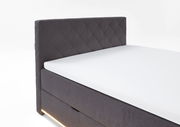 Seitliche Ansicht eines modernen Boxspringbetts mit grauem, gestepptem Kopfteil und integrierter Beleuchtung.