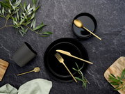 Schwarzes Geschirr- und Besteck-Arrangement mit goldfarbenem Messer, Gabel und Löffeln auf dunkler Steinoberfläche, mit grünen Zweigen und Holzbrett, Draufsicht (Flatlay)
