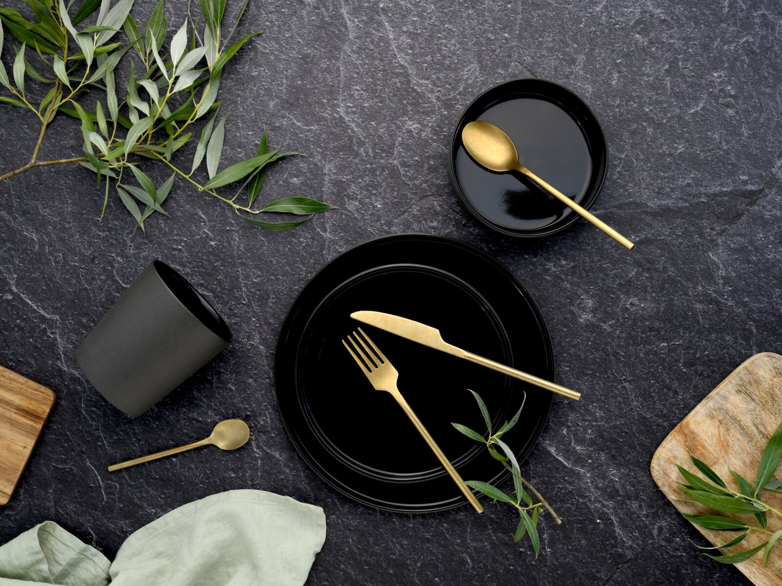 Schwarzes Geschirr- und Besteck-Arrangement mit goldfarbenem Messer, Gabel und Löffeln auf dunkler Steinoberfläche, mit grünen Zweigen und Holzbrett, Draufsicht (Flatlay)