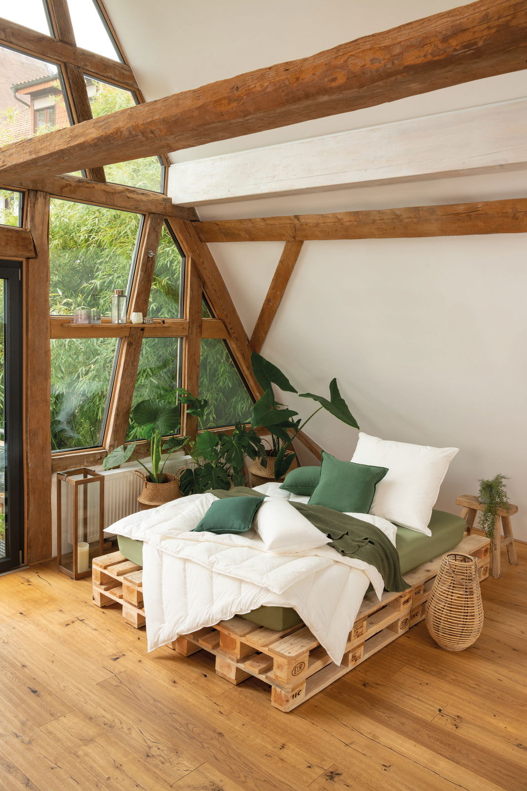 Gemütliches Schlafzimmer mit einem Solobett aus Paletten, umgeben von Pflanzen, in einem Raum mit großen Fenstern und Holzbalken, seitliche Perspektive.