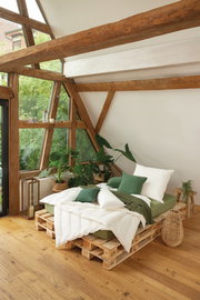 Gemütliches Schlafzimmer mit einem Solobett aus Paletten, umgeben von Pflanzen, in einem Raum mit großen Fenstern und Holzbalken, seitliche Perspektive.