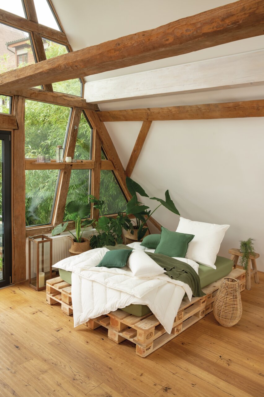 Gemütliches Schlafzimmer mit einem Solobett aus Paletten, umgeben von Pflanzen, in einem Raum mit großen Fenstern und Holzbalken, seitliche Perspektive.