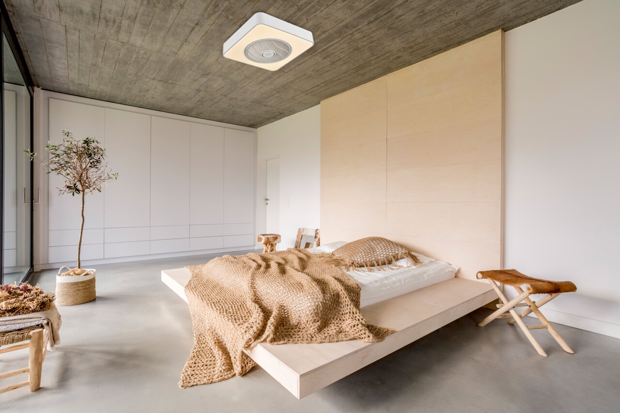 Modernes, minimalistisches Schlafzimmer mit niedrigem Holzplattformbett, beiger Strickdecke und Kissen, Beton-Decke mit Deckenleuchte, großer weißer Einbauschrank und Pflanze am Fenster; Perspektive: Weitwinkelaufnahme aus leicht erhöhter Ecke, Blick schr
