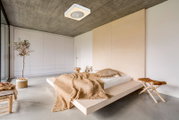 Modernes, minimalistisches Schlafzimmer mit niedrigem Holzplattformbett, beiger Strickdecke und Kissen, Beton-Decke mit Deckenleuchte, großer weißer Einbauschrank und Pflanze am Fenster; Perspektive: Weitwinkelaufnahme aus leicht erhöhter Ecke, Blick schr