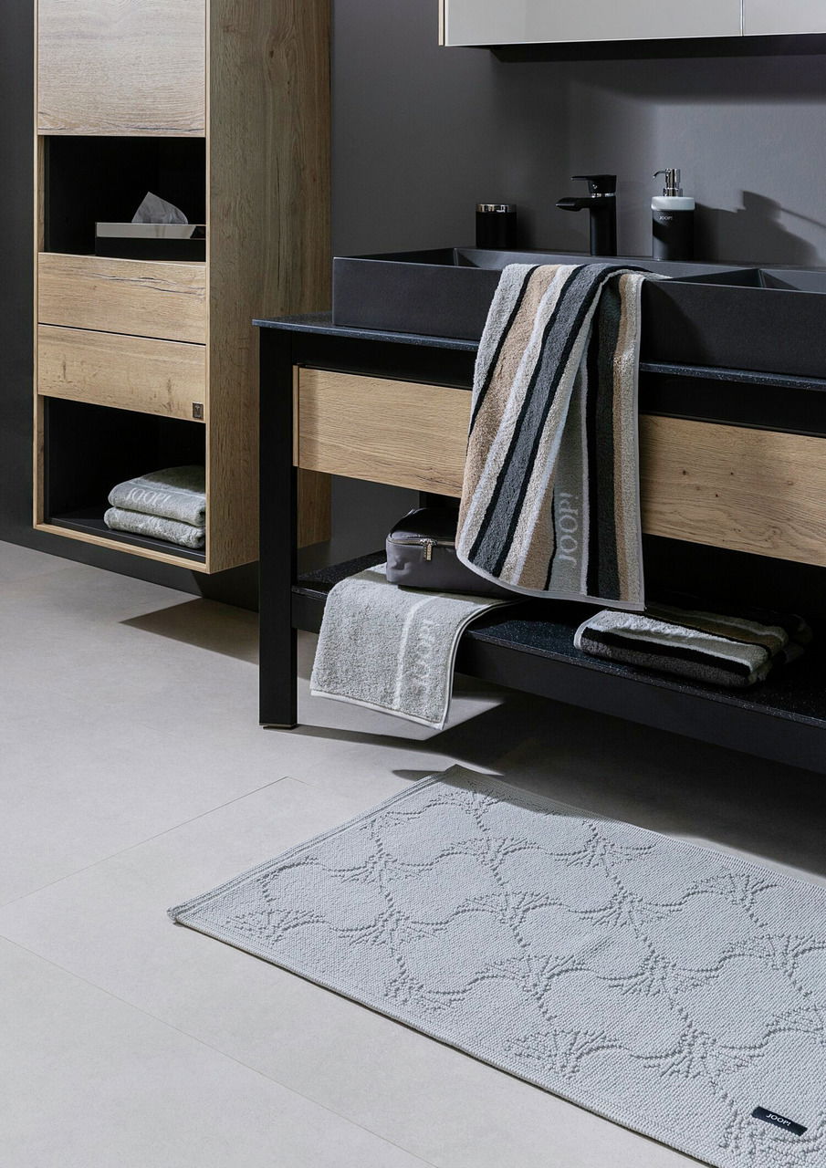 Moderne Badezimmeransicht mit schwarzer Mischbatterie und Waschbecken, umgeben von Holz- und Metallelementen, mit Handtüchern und Badteppich. Perspektive von schräg vorne.