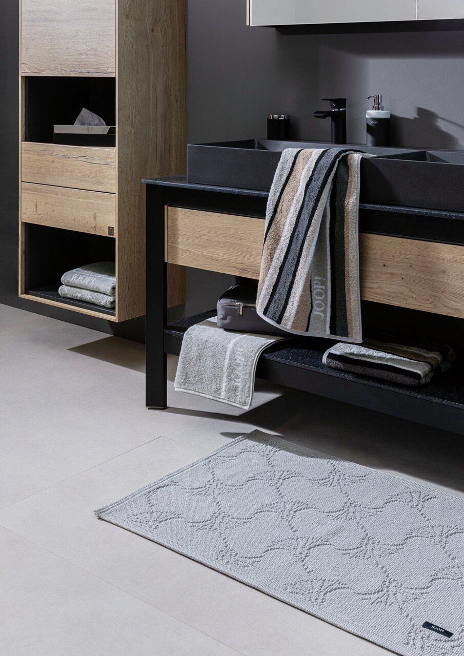 Moderne Badezimmeransicht mit schwarzer Mischbatterie und Waschbecken, umgeben von Holz- und Metallelementen, mit Handtüchern und Badteppich. Perspektive von schräg vorne.