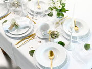 Schlicht gedeckter Festtisch in Weiß mit goldfarbenem Besteck, weißen Tellern und Schalen, Kristallgläsern, Kerzenständern und weiß-grünem Blumenarrangement auf weißer Tischdecke, aus schräger Vogelperspektive aufgenommen