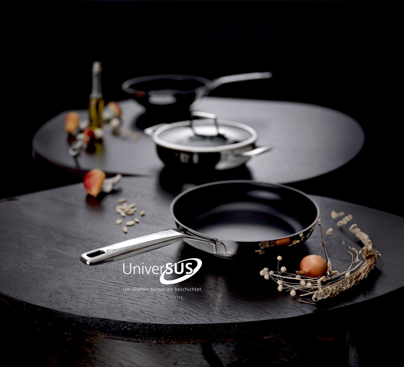 Schwarze Bratpfanne mit Edelstahlgriff auf dunklem Tisch, im Hintergrund Topf und weitere Pfanne, fotografiert aus leicht erhöhter Frontperspektive.