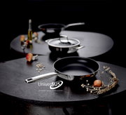 Schwarze Bratpfanne mit Edelstahlgriff auf dunklem Tisch, im Hintergrund Topf und weitere Pfanne, fotografiert aus leicht erhöhter Frontperspektive.