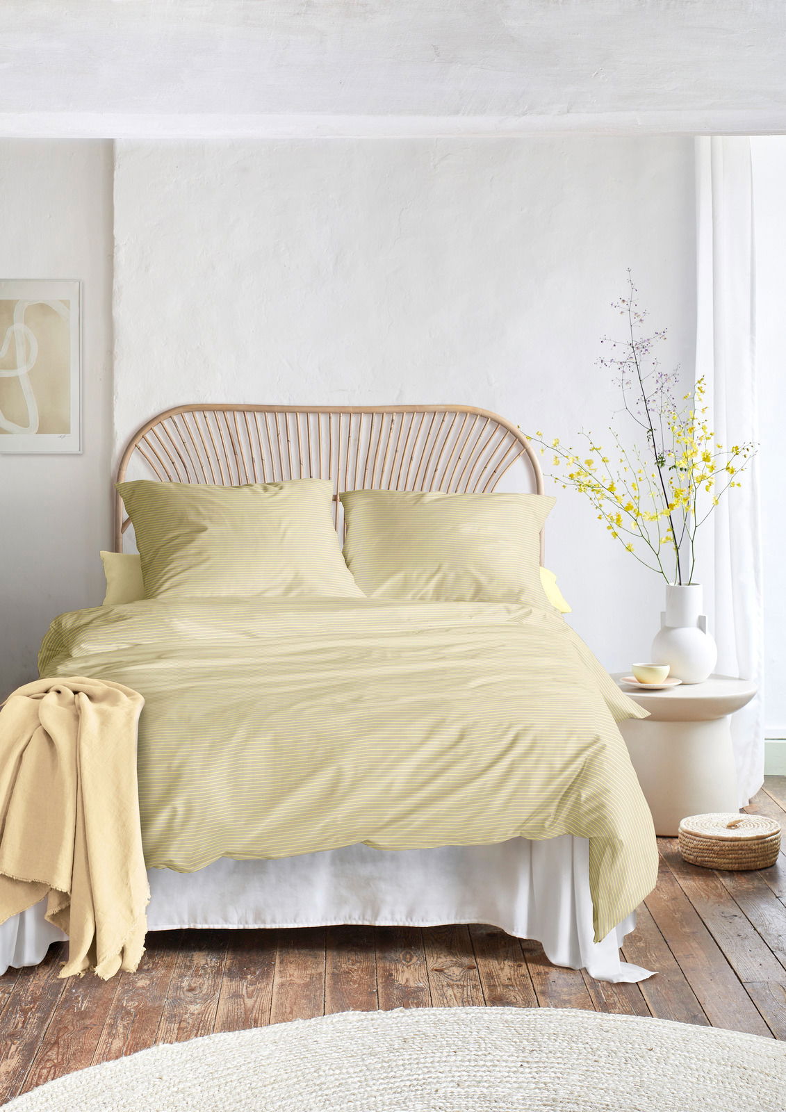 Bett mit gelber Bettwäsche in einem hellen Raum, frontal fotografiert, mit einem Rattan-Kopfteil und einem Beistelltisch mit Vase und Blumen.