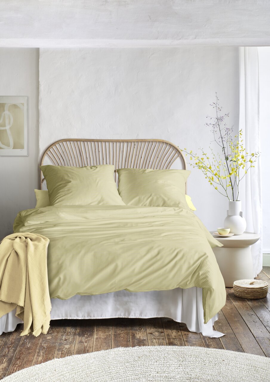 Bett mit gelber Bettwäsche in einem hellen Raum, frontal fotografiert, mit einem Rattan-Kopfteil und einem Beistelltisch mit Vase und Blumen.
