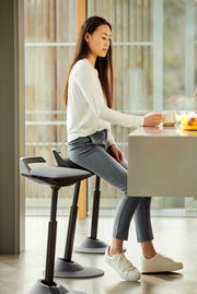 Frau sitzt auf einer Aeris Sitz-/Stehhilfe Muvman an einem Tisch, seitliche Perspektive.