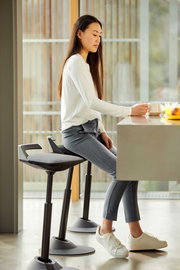 Frau sitzt auf einer Aeris Sitz-/Stehhilfe Muvman an einem Tisch, seitliche Perspektive.