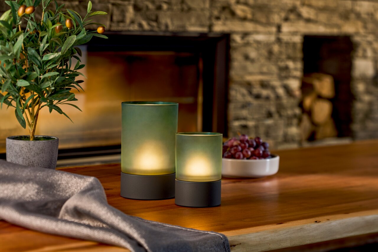 Zwei Akku-Windlichter in verschiedenen Größen auf einem Holztisch, umgeben von einer Pflanze, einer Schale mit Trauben und einem Kamin im Hintergrund. Seitliche Perspektive.