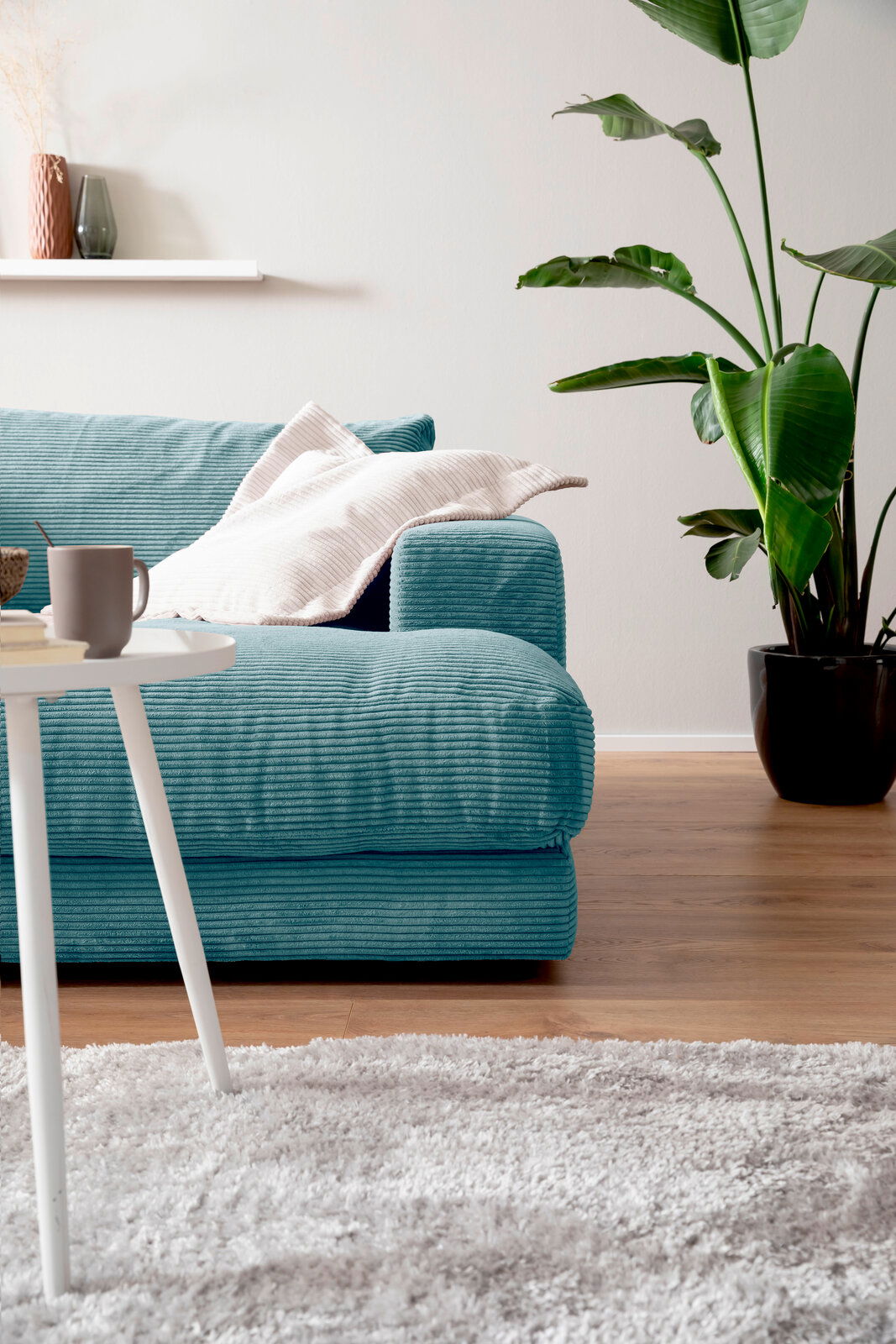 Ein modernes, kleines Megasofa in Türkis mit Kissen, im Vordergrund ein weißer Tisch und im Hintergrund eine grüne Pflanze, seitliche Perspektive.