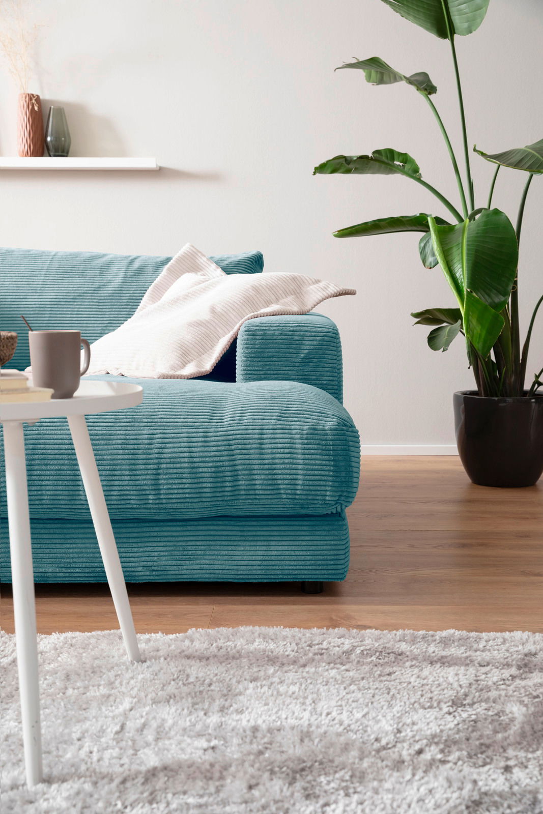 Ein modernes, kleines Megasofa in Türkis mit Kissen, im Vordergrund ein weißer Tisch und im Hintergrund eine grüne Pflanze, seitliche Perspektive.