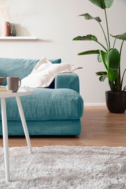 Ein modernes, kleines Megasofa in Türkis mit Kissen, im Vordergrund ein weißer Tisch und im Hintergrund eine grüne Pflanze, seitliche Perspektive.