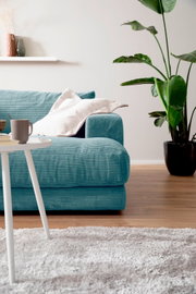 Een moderne, kleine megabank in turkoois met kussens, op de voorgrond een witte tafel en op de achtergrond een groene plant, gezien vanuit een zijdelingse hoek.
