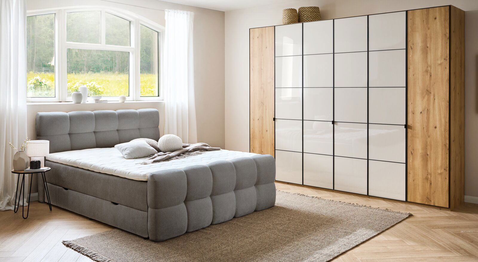 Graues Boxbett in einem hellen Schlafzimmer mit großem Fenster, neben einem modernen Kleiderschrank. Perspektive von vorne links.