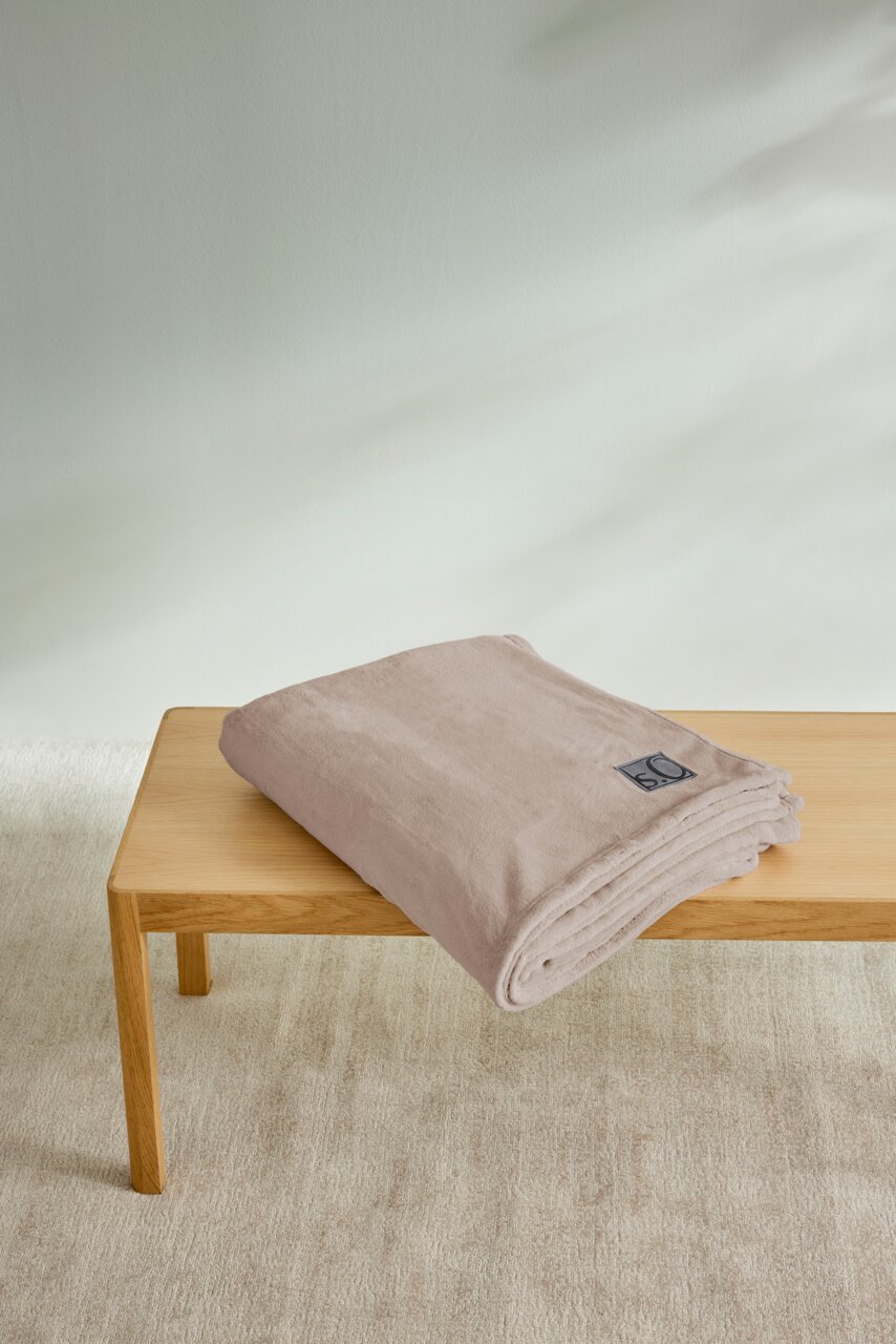 Beige Kuscheldecke von s.Oliver, gefaltet auf einem Holztisch liegend, aus seitlicher Perspektive fotografiert.