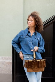 Frau in Denim-Outfit hält eine braune Wildleder-Handtasche mit Fransen und hellem Griff, seitliche Halbprofil-Ansicht in leichtem Untersichtwinkel.