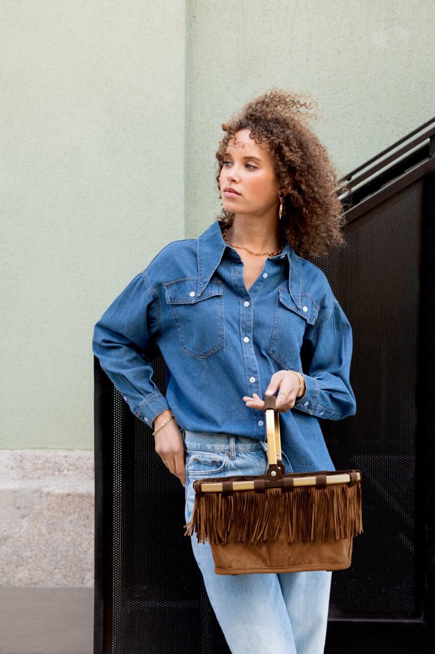 Frau in Denim-Outfit hält eine braune Wildleder-Handtasche mit Fransen und hellem Griff, seitliche Halbprofil-Ansicht in leichtem Untersichtwinkel.